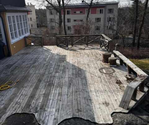 Grå träaltan formad runt en berghäll framför ett gult hus före rengöring.